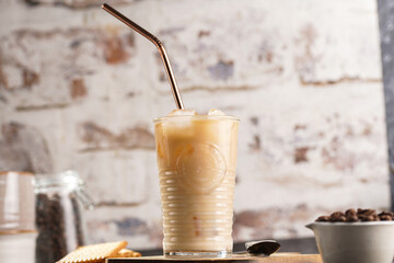 A glass with ice cubes and milk coffee on a wooden board, with brass straw