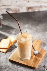 A glass with ice cubes and milk coffee on a wooden board, with brass straw