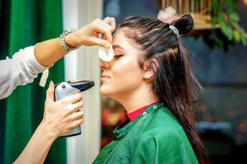 Side view of a make-up artist using aerograph making an airbrush makeup foundation on a female face...