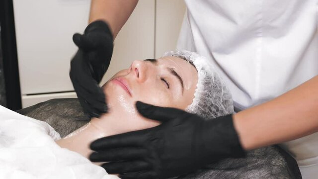 beauty therapy. Cosmetology skin care. beautician in black medical gloves performs cosmetic skincare procedure, applies cleansing foam or emulsion to female client's face. close-up. anti aging