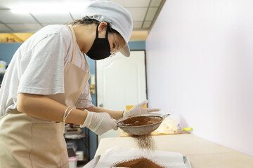 Happy young Asian woman with protective face mask, apron, glove, hat is smiling while using flour...