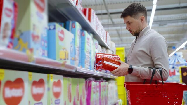 Young Father Is Buying Diapers In Supermarket, Choosing Best Good For His Child