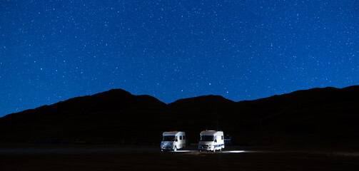 Campers parked in a caravan parking area sleeping on a starry night in the mountain. Summer tourism with RV in a blue night sky with stars. Best option for travel. Panorama motorhomes and camping car. - obrazy, fototapety, plakaty