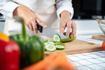 Asian housewife wearing apron and using knife to slice cucumber and tomato on chopping board