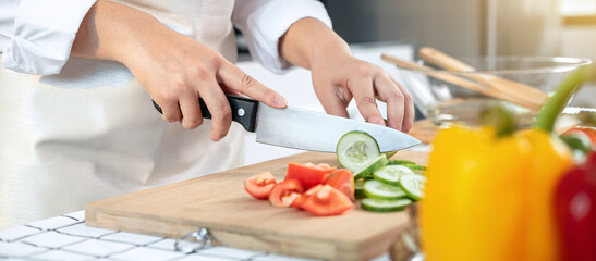 Close up of asian housewife wearing apron and using knife to slice cucumber and tomato on chopping board