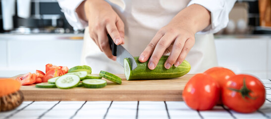 Close up of asian housewife wearing apron and using knife to slice cucumber and tomato on chopping board