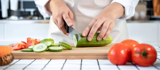 Fotobehang Close up of asian housewife wearing apron and using knife to slice cucumber and tomato on chopping board © Freedomz