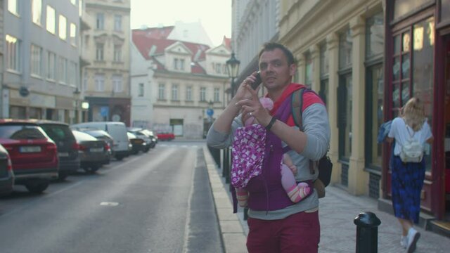 A man's dad is holding a small child in a sling. He Uses the phone, makes a call to the service. Waiting for a taxi car in the city.