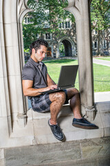 Dressing in a gray shirt, pattern shorts and black leather sneakers, a young handsome student is sitting on a old fashion window frame and study on a computer in a university campus