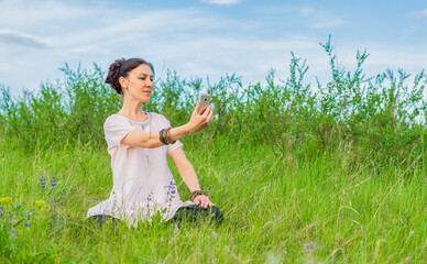 A beautiful young woman sits on a green lawn in a lotus position, holds a phone in her hand and looks at it. Outdoor recreation.