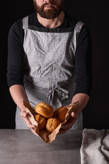 A man baker with a beard in a gray apron stands against a black background and holds, breaks, cuts off delicious, crispy bread, rolls, baguette