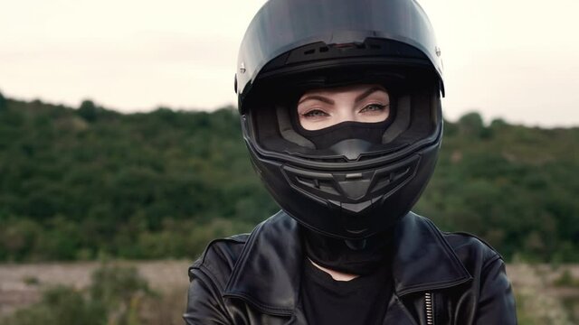 Young stylish motorcyclist woman in motorcycle helmet and leather jacket. Sexy biker lady outdoors on nature background. Trip, cafe racers, speed, freedom concept.