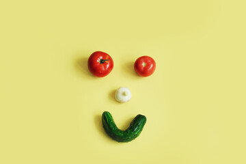 Vegetables depicting a vegetable face, tomato eyes, garlic nose, cucumber smile on a yellow background, top view, copy space.