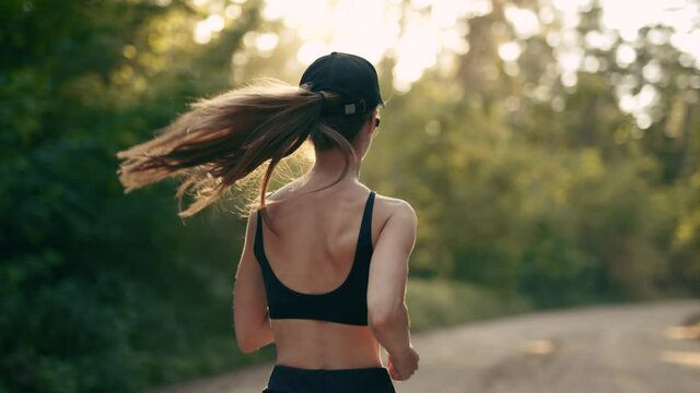 A young woman runner is listening to music in earphones and training in summer forest. Close up of fitness girl is jogging outdoor. Concept of workouts running and healthy lifestyle. Back view.