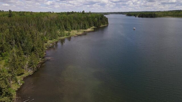 4K flyover of beautiful, rippling, blue lake and treed shoreline on a cloudy day. Pontoon boat can be seen on lake. Lake Itasca, MN. 