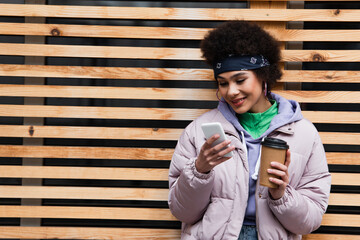 Fototapeta premium Positive african american woman using mobile phone and holding paper cup near fence