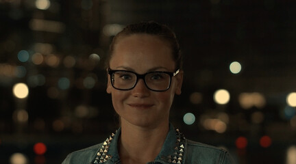 Portrait of beautiful positive young girl in casual clothes and glasses posing on the background of glowing nighttime buildings in the metropolis holding smartphone. Concept of tourist navigators apps