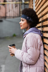 Fototapeta na wymiar Side view of african american woman in bandana and jacket holding paper cup near wooden fence