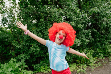 Child clown in a red wig and nose stands in the open air and smiles. April 1. Day of Laughter. Birthday.