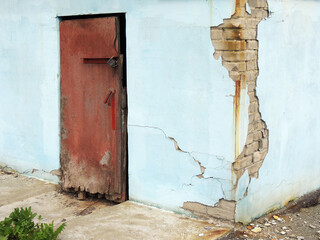 A burgundy, old, closed door with a lock and a handle against a blue wall. The brick wall is covered with blue plaster. The plaster is falling off the wall. Bricks and cracks are visible on the wall. 