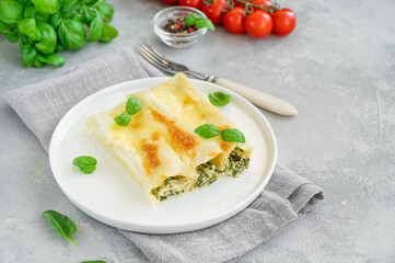 Traditional Italian cannelloni with ricotta and spinach in bechamel sauce with fresh basil leaves on a white plate on a gray concrete background. Vegetarian dish. Copy space.
