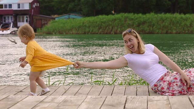 Young caucasian mother holding her little daughter on the dress and funny fallen, playing on the wooden pier by river. Side view. Slow motion. The concept of children's education.
