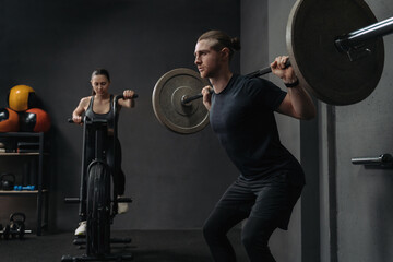 Fototapeta na wymiar Couple have crossfit training at gym. Attractive fit woman is working out on exercise bike, while muscular man training with barbell. Functional and circuit training concept