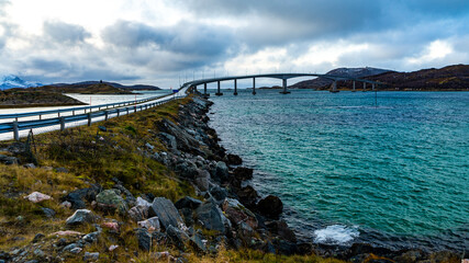Brücke zur Insel Sommaroy in Troms, in der Nähe von Tromsö, Norwegen, am Atlantik. grün blaues Meer bei stürmischem Wetter, Steine und Sand am Ufer, mit Tang und Gras - obrazy, fototapety, plakaty