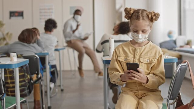 Medium slowmo of teenage red-haired school girl in face mask playing games on smartphone sitting in front of camera with her back to classmates and teacher having lesson