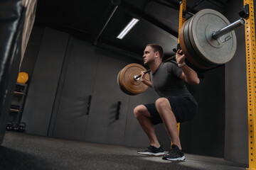 Obraz na płótnie Canvas Muscular caucasian man lifting weights and doing back squat in gym. Crossfit athlete is holding a heavyweight barbell on the shoulder behind the neck while squatting. Fitness and work out