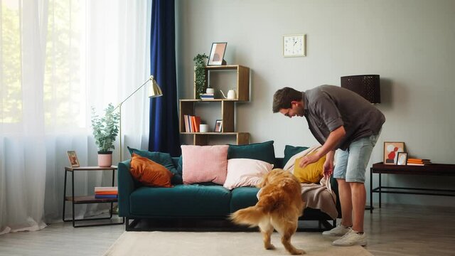 Young man training his dog, throwing small ball to obedient golden retriever in living-room. Energetic puppy catching yellow toy. Playing and spending time together with lovely pet at home.