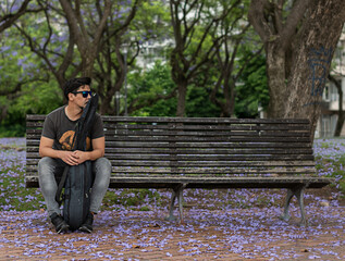 guitarist man sitting in a bank isolated in a park with falling flowers on the floor waiting  thoughtful with guitar inside the case