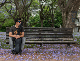guitarist man sitting in a bank isolated in a park with falling flowers on the floor waiting ...