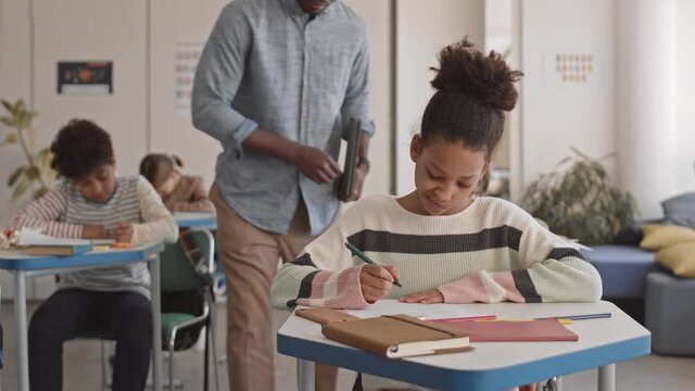 Medium shot of modern young male Afro American teacher walking along row in classroom pointing at piece of paper lying on students desks helping kids while they doing important test