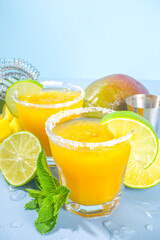 Cold summer cocktail, mango margaritas with tequila, salt lime slices, crushed ice and mint. Seasonal refreshing drink, on bright blue sun lighted background