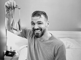 Mature happy man with chronic breathing issues considers using CPAP machine sitting on the bed in bedroom . Healthcare, CPAP, snoring concept