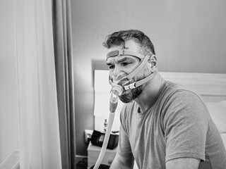 Unhappy shocked man with chronic breathing issues surprised by using  CPAP machine sitting on the bed in bedroom. Healthcare, CPAP, Obstructive sleep apnea therapy, snoring concept