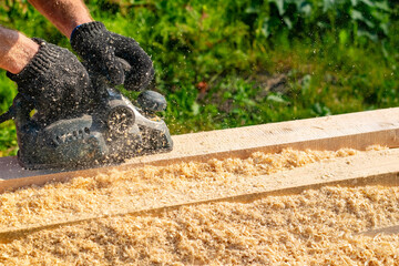 the hands of a person, a worker, a carpenter hold an electric plane, with which a builder planks a long board on the street and shavings and dust fly