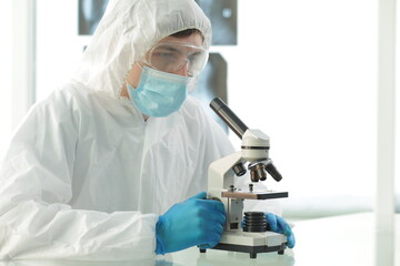 Doctor in a protective medical suit wearing a mask wearing blue gloves in a laboratory looks through a microscope