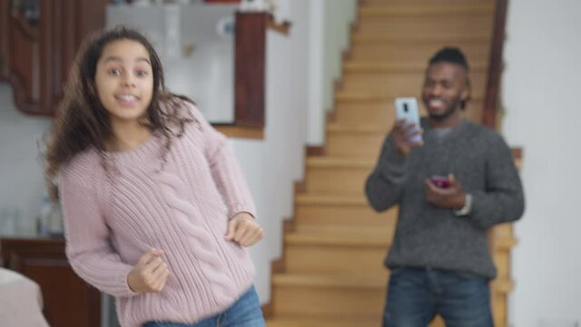Medium shot of carefree cheerful African American teen girl having fun dancing in living room with smiling blurred man filming on smartphone standing at background. Positive family indoors