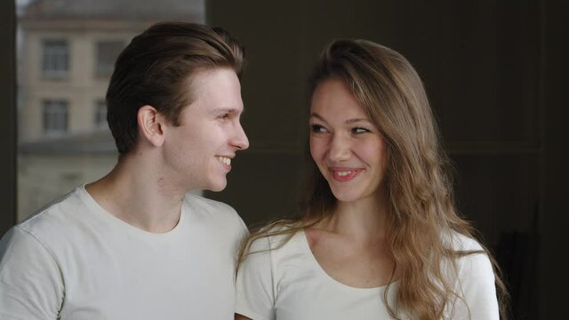 Caucasian young married couple lovers of millennials stand together indoors boyfriend kissing girlfriend on cheek woman wife girl smiling embarrassed after kiss husband man expresses love tenderness