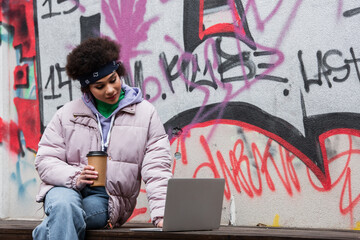 African american woman holding coffee to go and using laptop near graffiti outdoors