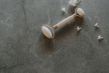 Gua sha jade facial roller - lifting, anti-wrinkle, anti-aging  tool. White beauty roller, massager and lilac flowers on a grey stone background.