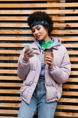 African american woman with coffee to go using cellphone near wooden fence