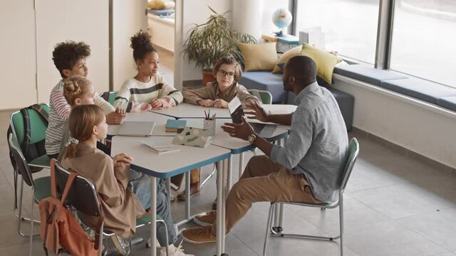 High-angle slowmo of multiethnic 5 or 6 grade students and young male Afro American teacher in casualwear sitting in circle in bright classroom having funny interesting Geography lesson
