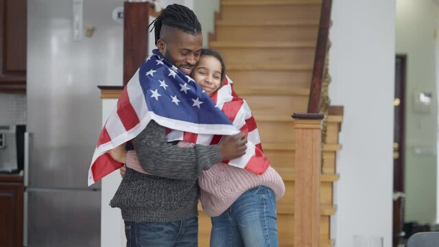 Happy African American adult man and teen girl posing with USA flag smiling and hugging. Portrait of cheerful proud father and daughter holding national symbol looking at camera. Pride and family