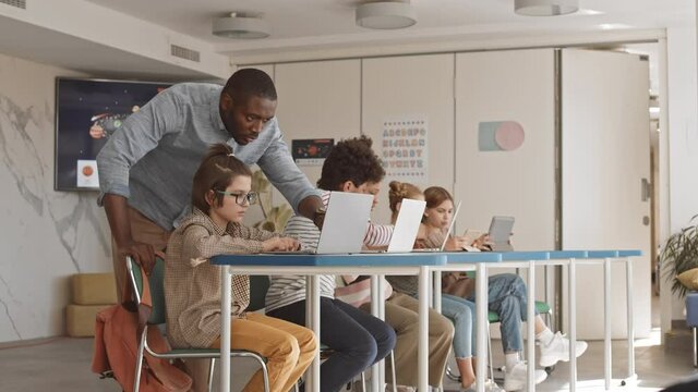 Slowmo of Afro-American male school teacher helping 5 or 6 grade multiethnic students with studies on laptop in modern IT classroom