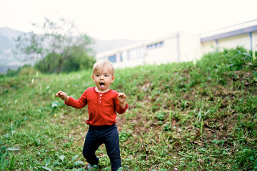 Kid walks on the green grass along the hillside against the backdrop of buildings