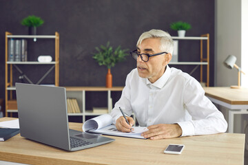 Busy senior male worker sitting at laptop making notes while working in modern coworking center. Concentrated serious businessman sitting at the table checks paper and electronic documents.