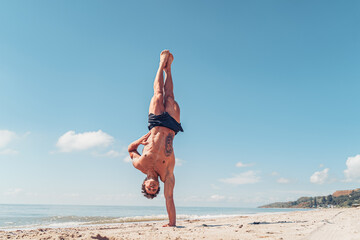 Muscular bodybuilder fitness man with a naked torso stands on one arm on the beach